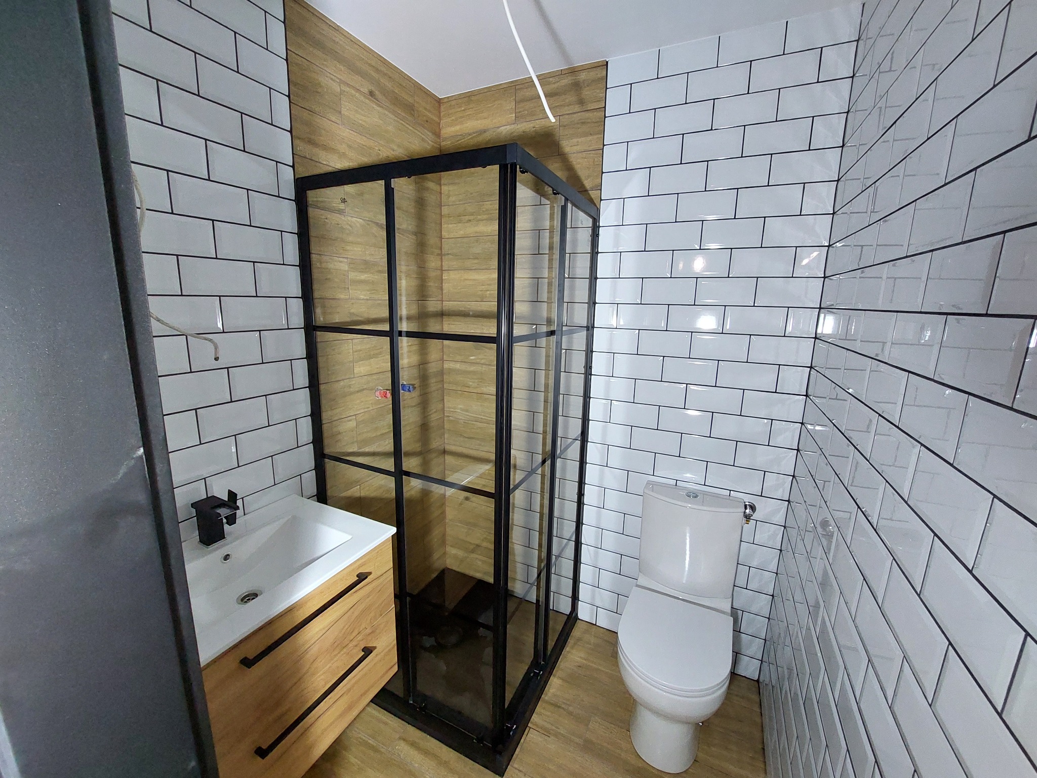 Nowoczesna łazienka z kabiną prysznicową typu walk-in z czarnymi profilami, umywalką z szafką z jasnego drewna i białą toaletą, wykończona białymi płytkami imitującymi cegłę i panelami...