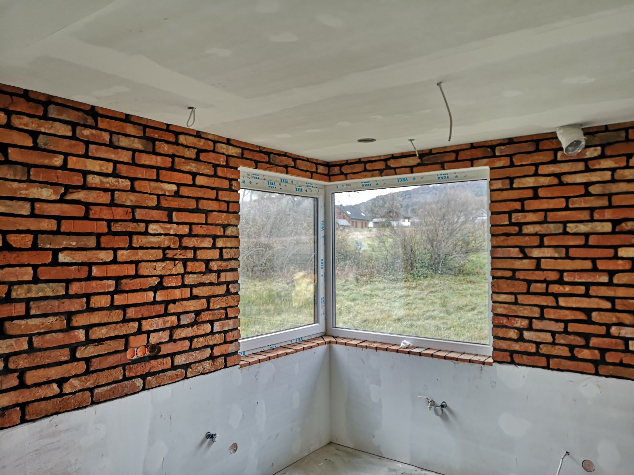 Wnętrze w trakcie wykańczania z ceglaną ścianą, dwoma oknami narożnymi z widokiem na zewnątrz, surowym sufitem i ścianami przygotowanymi do malowania.