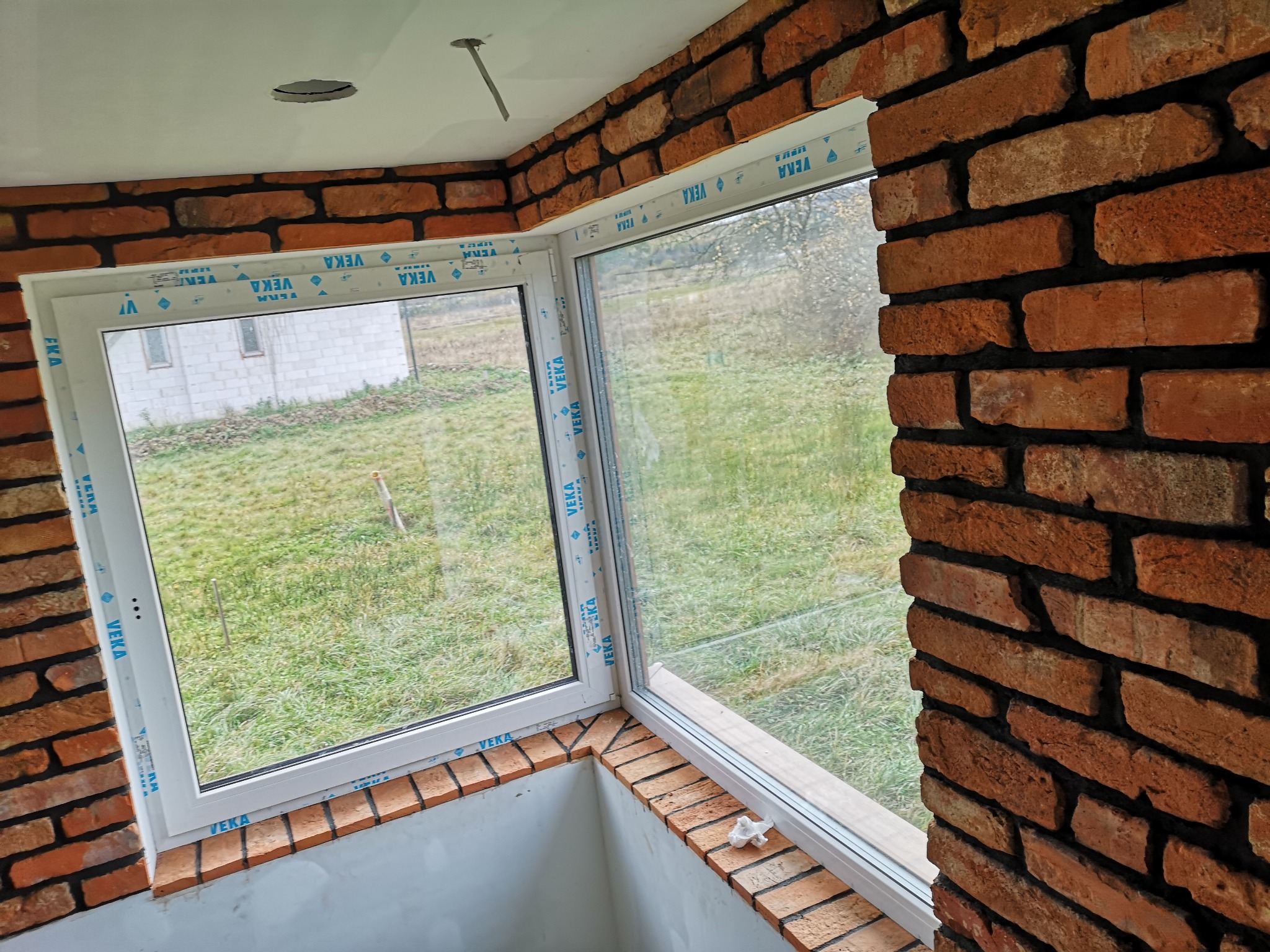 Wnętrze z oknami PCV marki Veka, obłożone czerwoną cegłą klinkierową, widok na zewnątrz na trawnik i budynek gospodarczy, surowy stan wykończenia.