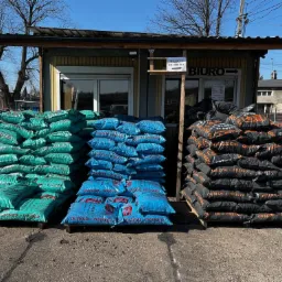 Skladowane worki z opałem: zielone, niebieskie i czarne, ułożone na paletach przed budynkiem biura z szyldem 'Wejście do Biura'.