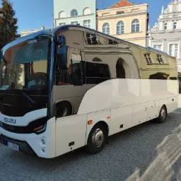 Biały autokar Isuzu na brukowanym placu miejskim, w szybach odbijają się kolorowe kamienice.