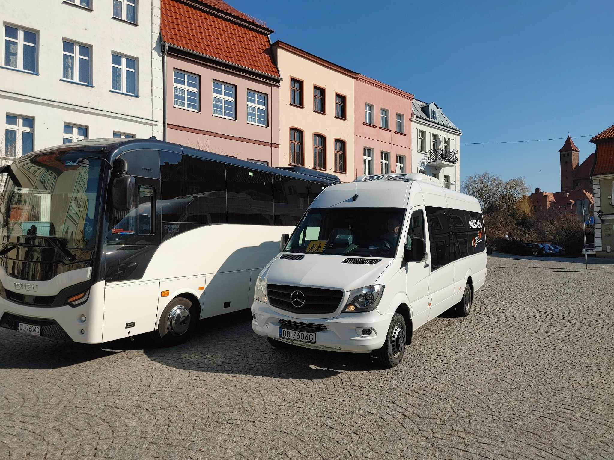 Biały bus Mercedes-Benz i autokar Isuzu na brukowanym placu miejskim, w tle kolorowe kamienice i wieża zamkowa.