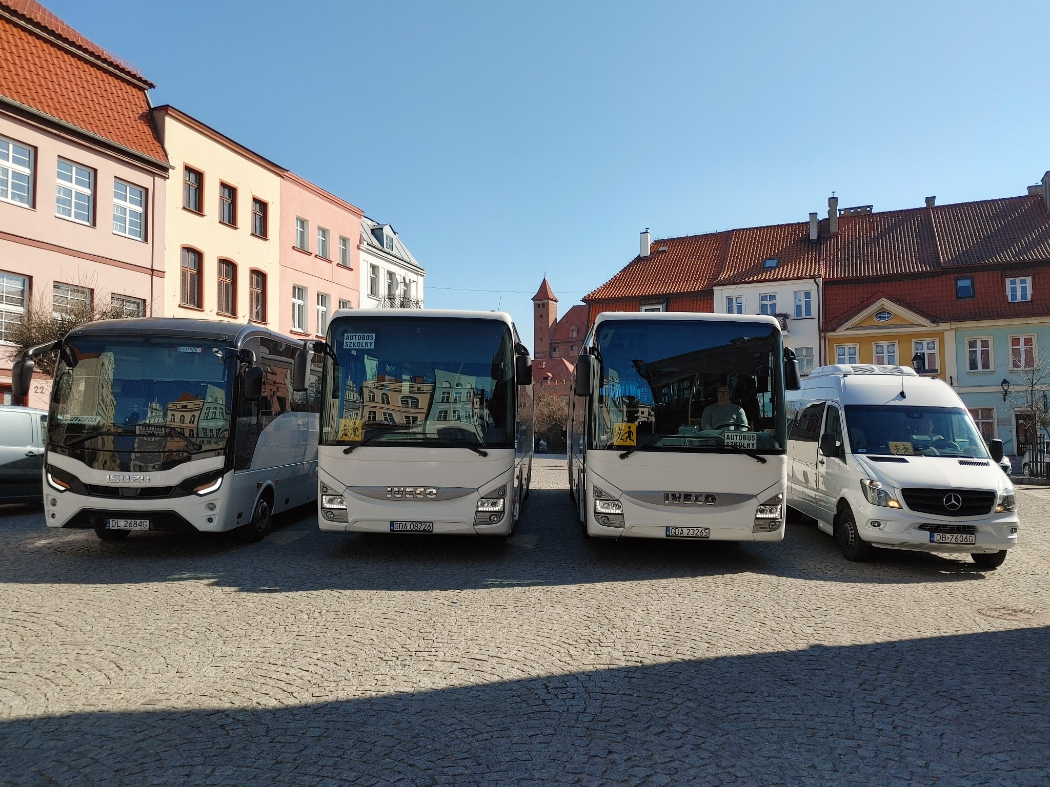 Flota pojazdów transportowych: autokar Isuzu, dwa busy Iveco z oznaczeniem 'Autobus Szkolny' i biały van Mercedes, ustawione na brukowanym placu w historycznym centrum miasta.