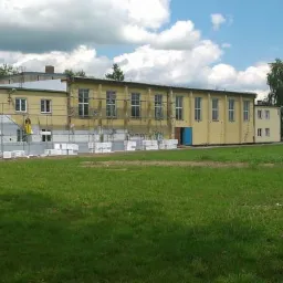 Budynek w trakcie prac elewacyjnych, widoczne rusztowania, częściowo ocieplona ściana i materiały budowlane na trawniku.