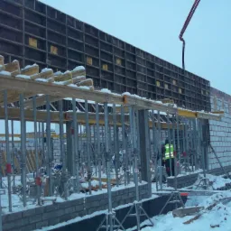 Budowa fundamentów w zimowych warunkach: szalunek zbrojony wsparty metalowymi stemplami, częściowo obłożony bloczkami, z widoczną pompą do betonu i pracownikiem w odblaskowej kamizelce.