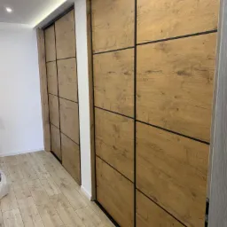 Dwie szafy wnękowe z przesuwnymi drzwiami o drewnianej fakturze i czarnych podziałach, widoczne na tle parkietu i białej ściany.