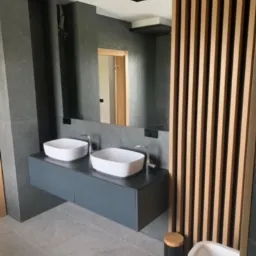 Nowoczesna łazienka z dwoma umywalkami nablatowymi o kwadratowym kształcie, zamontowanymi na szarej, wiszącej szafce. Na ścianie duże lustro z podświetleniem. Drewniane lamele tworzą dekoracyjną...