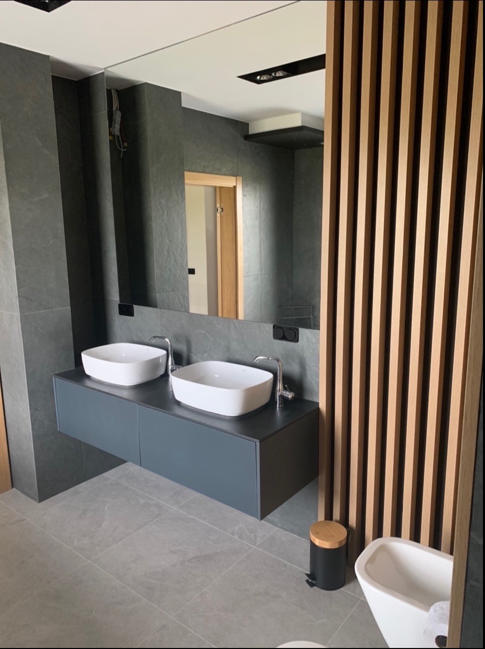 Nowoczesna łazienka z dwoma białymi umywalkami nablatowymi na szarej, wiszącej szafce, szare płytki na ścianach i podłodze, drewniana ścianka działowa z pionowych listew oraz duże lustro. Widoczny...