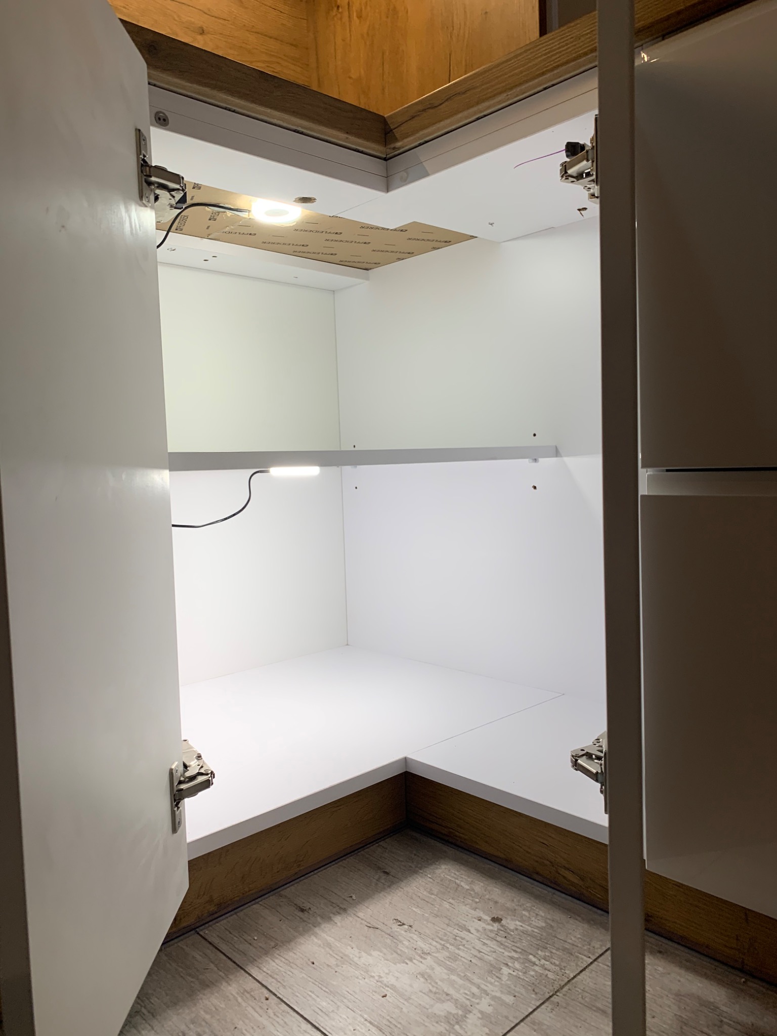 Wnętrze narożnej szafki z białymi półkami i oświetleniem LED, widoczne zawiasy i fragmenty drewnianej obudowy.
