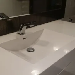 Biała, nowoczesna umywalka wpuszczona w blat z minimalistyczną baterią, odbicie grzejnika dekoracyjnego w lustrze nad umywalką.