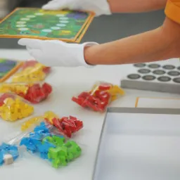 Ręce w białych rękawiczkach układają planszę gry obok zapakowanych woreczków z kolorowymi pionkami i pustego pudełka.