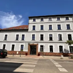 Odnowiona kamienica z szarą elewacją, ciemnymi oknami z białymi obramowaniami, widoczna z poziomu ulicy z przejściem dla pieszych i fragmentami sąsiednich budynków.