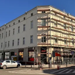 Kamienica w trakcie malowania elewacji, widoczne rusztowania, jasna fasada i błękitne niebo w słoneczny dzień.