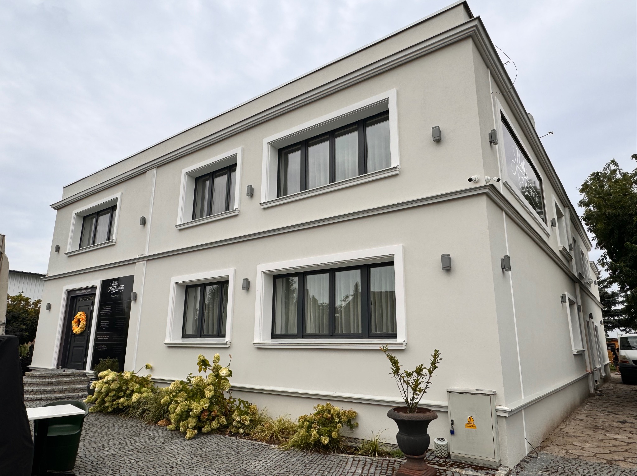 Dwupiętrowy budynek z jasną elewacją, czarnymi ramami okiennymi, oświetleniem zewnętrznym i dekoracyjnym wieńcem na drzwiach wejściowych, otoczony roślinnością i brukowanym chodnikiem.