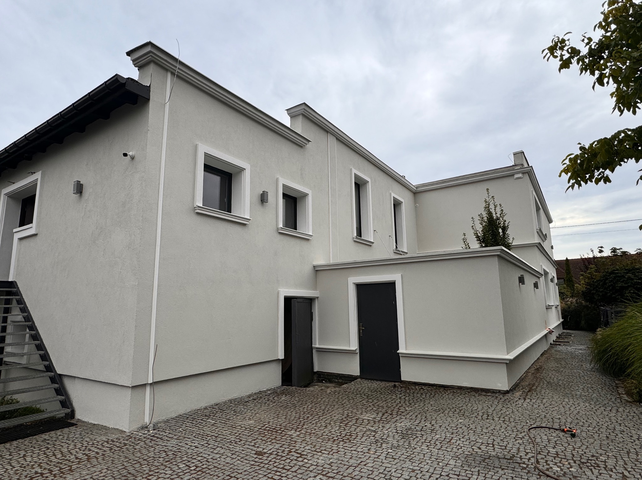 Dwupiętrowy budynek z jasnoszarą elewacją, widoczne okna z białymi obramowaniami, czarne drzwi wejściowe i schody zewnętrzne po lewej stronie, kostka brukowa na podjeździe.