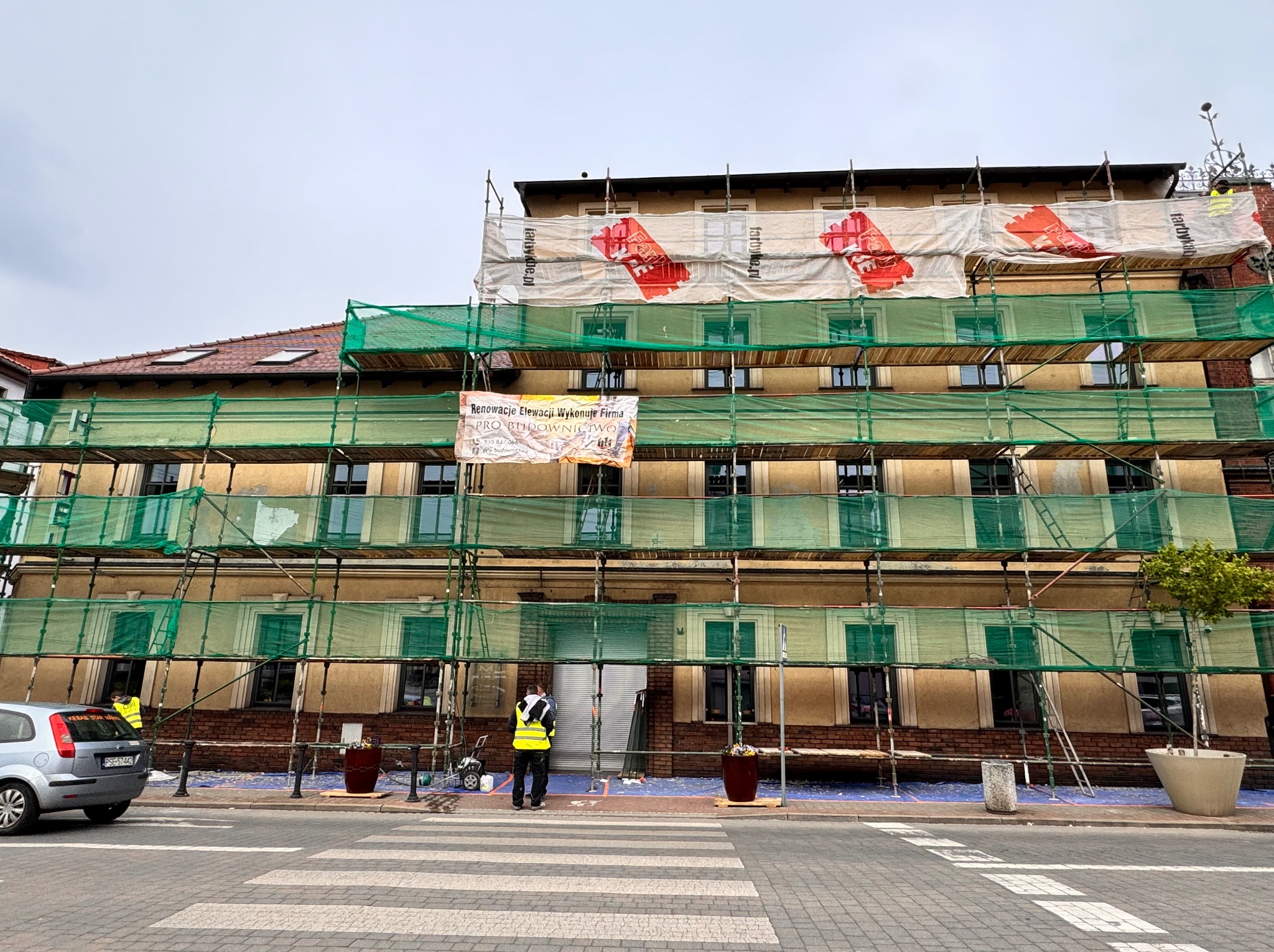 Budynek Hotelu Metropol w Śremie w trakcie renowacji elewacji, widoczne rusztowania z zieloną siatką, banery reklamowe farb i ekipa remontowa w żółtych kamizelkach na chodniku.