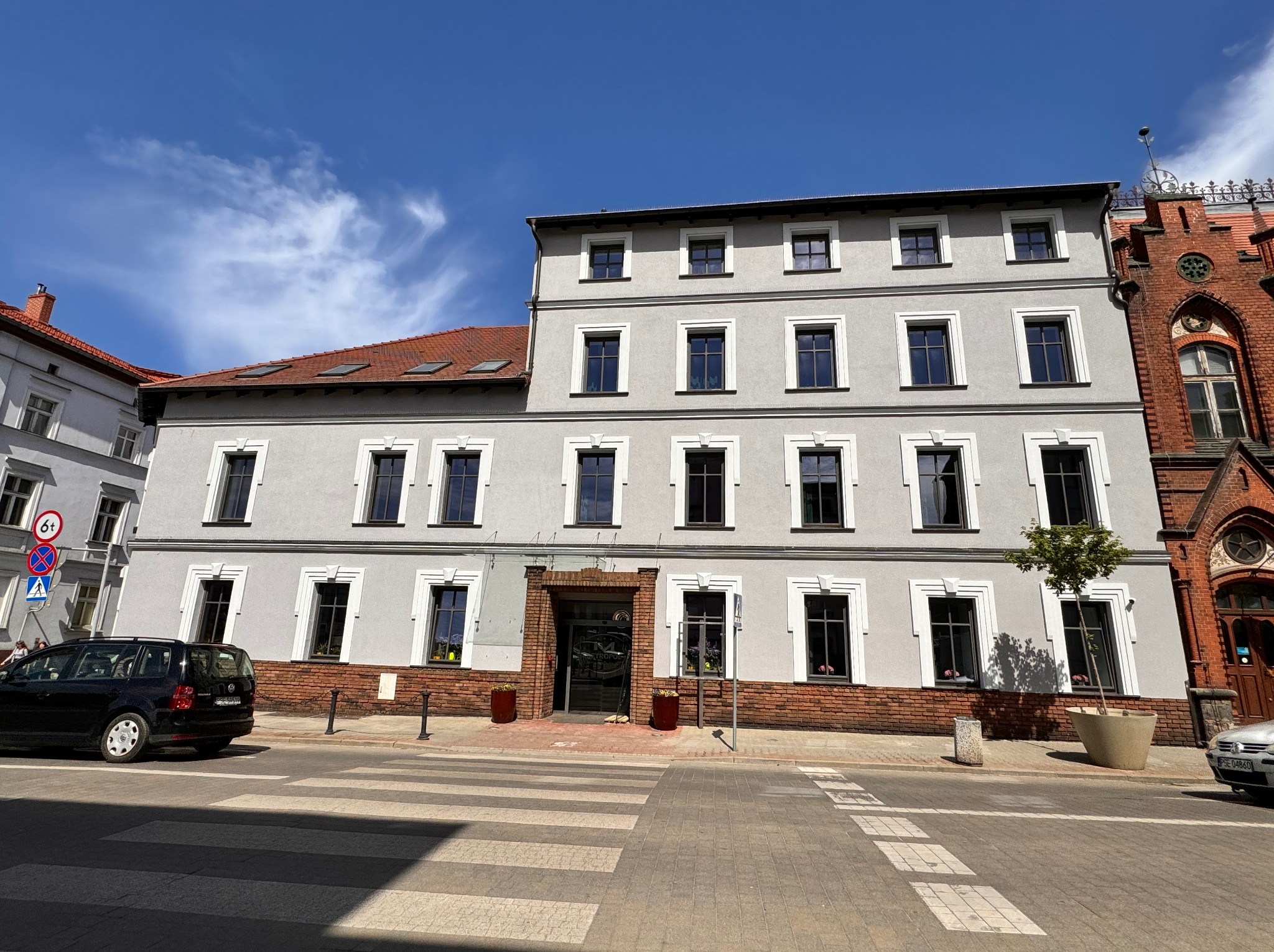 Odnowiona kamienica z szarą elewacją, ciemnymi oknami z białymi obramowaniami, widoczna z poziomu ulicy z przejściem dla pieszych i fragmentami sąsiednich budynków.