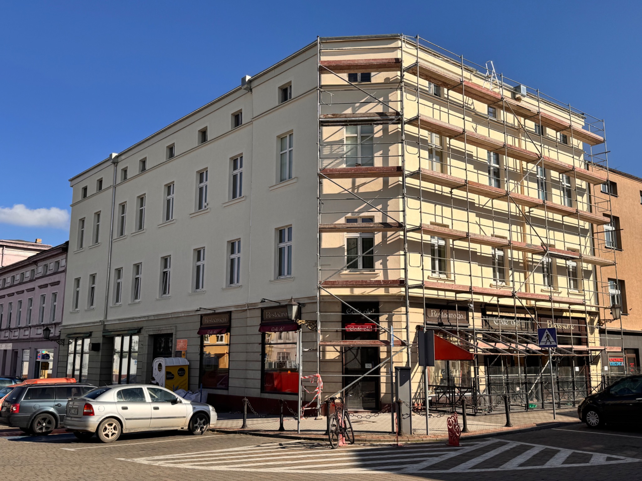 Kamienica w trakcie malowania elewacji, widoczne rusztowania, jasna fasada i błękitne niebo w słoneczny dzień.