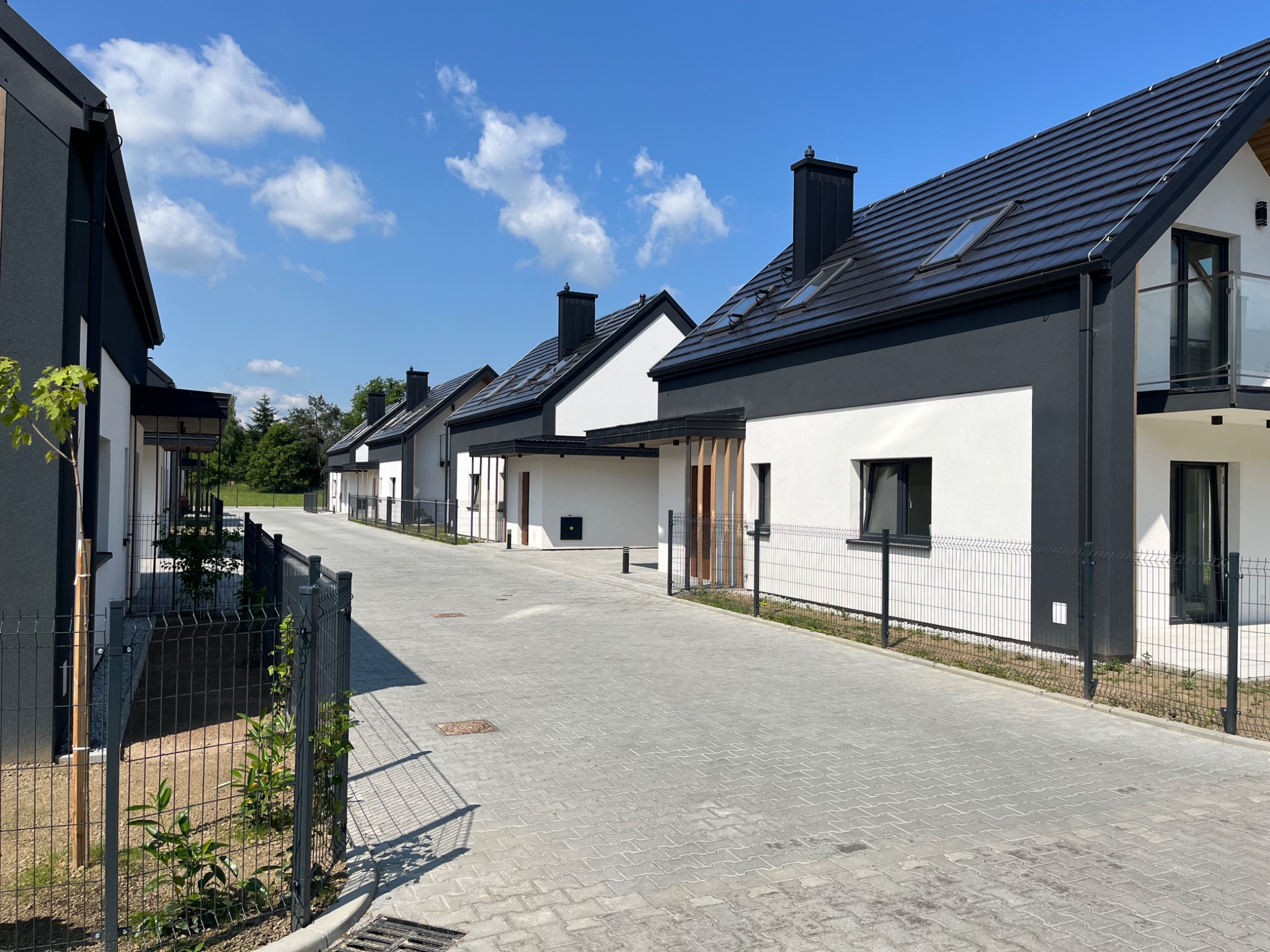 Nowoczesne osiedle domów jednorodzinnych w zabudowie szeregowej, z czarno-białą elewacją, czarną dachówką i ogrodzeniem panelowym, widok z poziomu ulicy w słoneczny dzień.