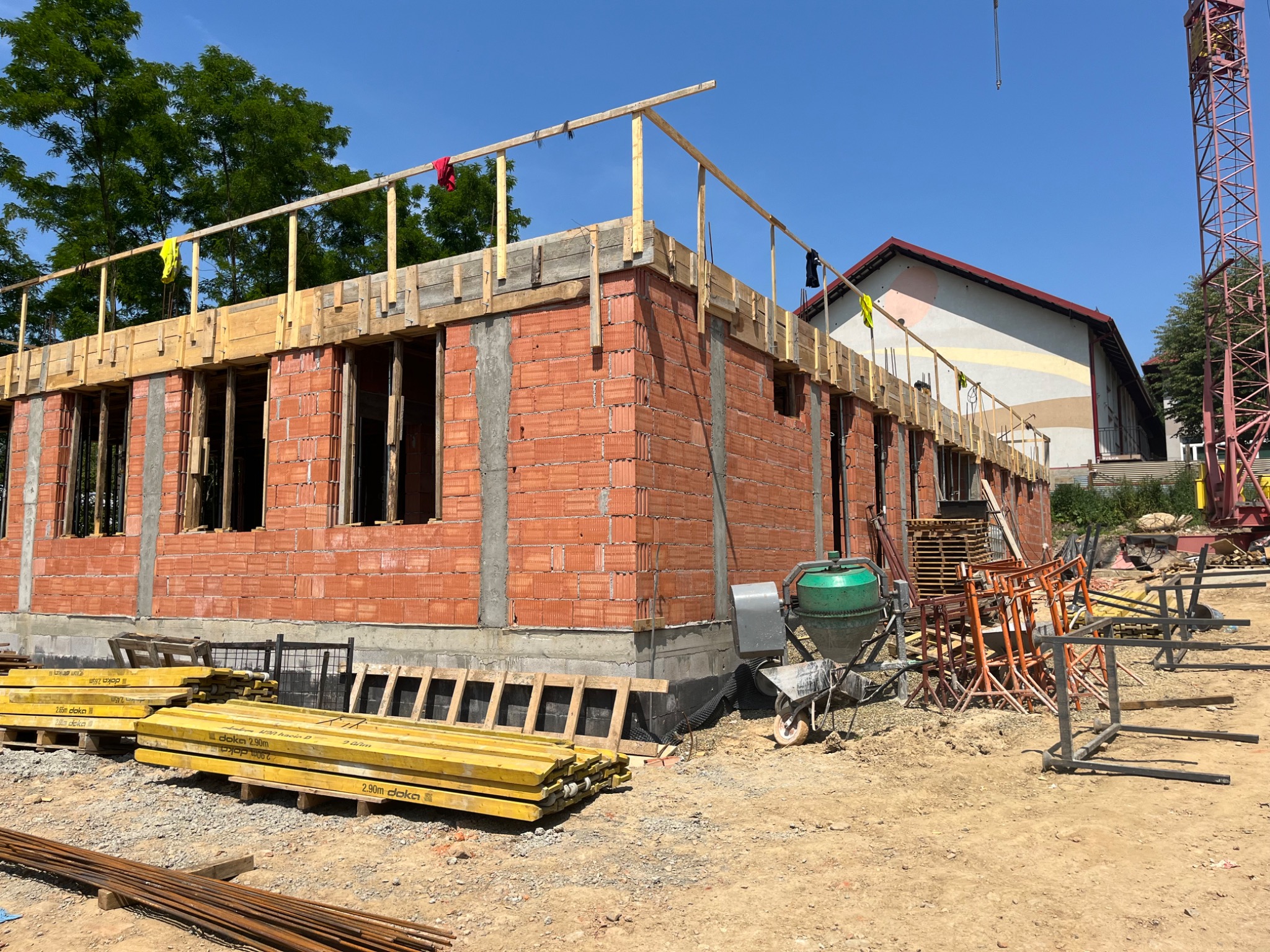 Budowa domu jednorodzinnego w trakcie wznoszenia murów z czerwonej cegły, widoczne szalunki i podpory, na pierwszym planie materiały budowlane oraz betoniarka.