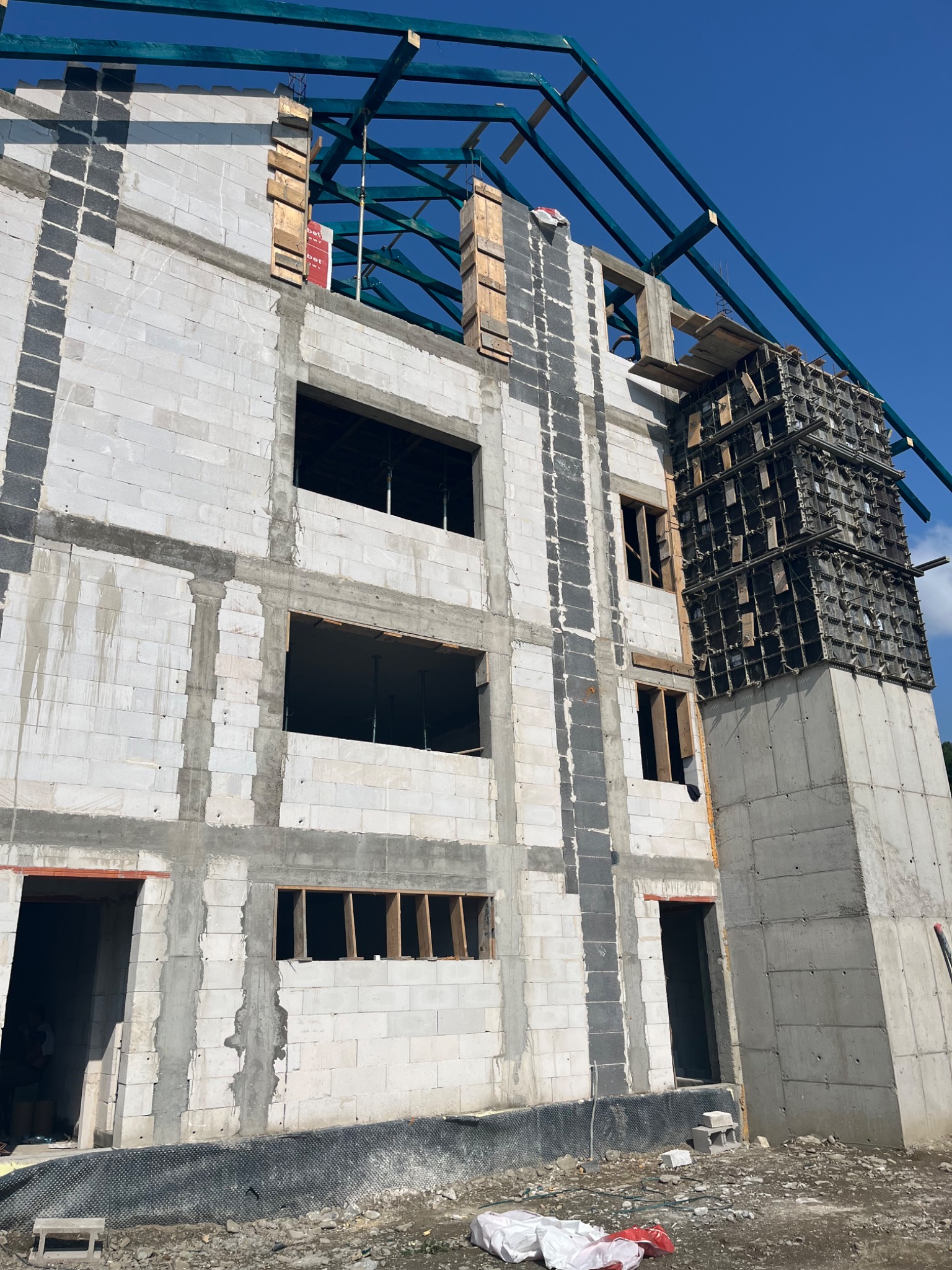 Surowy stan budynku wielopiętrowego z betonu komórkowego i szalunków kolumnowych, widoczne niebieskie elementy konstrukcji dachu i błękitne niebo w tle.