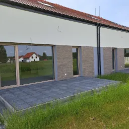 Elewacja szeregowca z dużymi oknami z drewnianą ramą i wyjściem na taras z szarej kostki, widok z poziomu trawy.