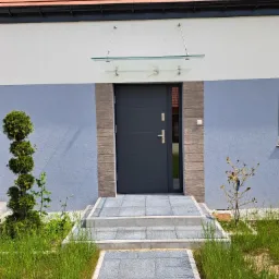 Nowoczesne, antracytowe drzwi wejściowe z przeszklonym panelem bocznym, osłonięte szklanym daszkiem, prowadzące po stopniach z szarej kostki, otoczone zielenią.