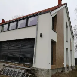 Nowoczesny dom z oknami z szarymi roletami zewnętrznymi, elewacją w kolorze złamanej bieli i elementami dekoracyjnymi z paneli imitujących drewno, widoczne rusztowanie i materiały budowlane.