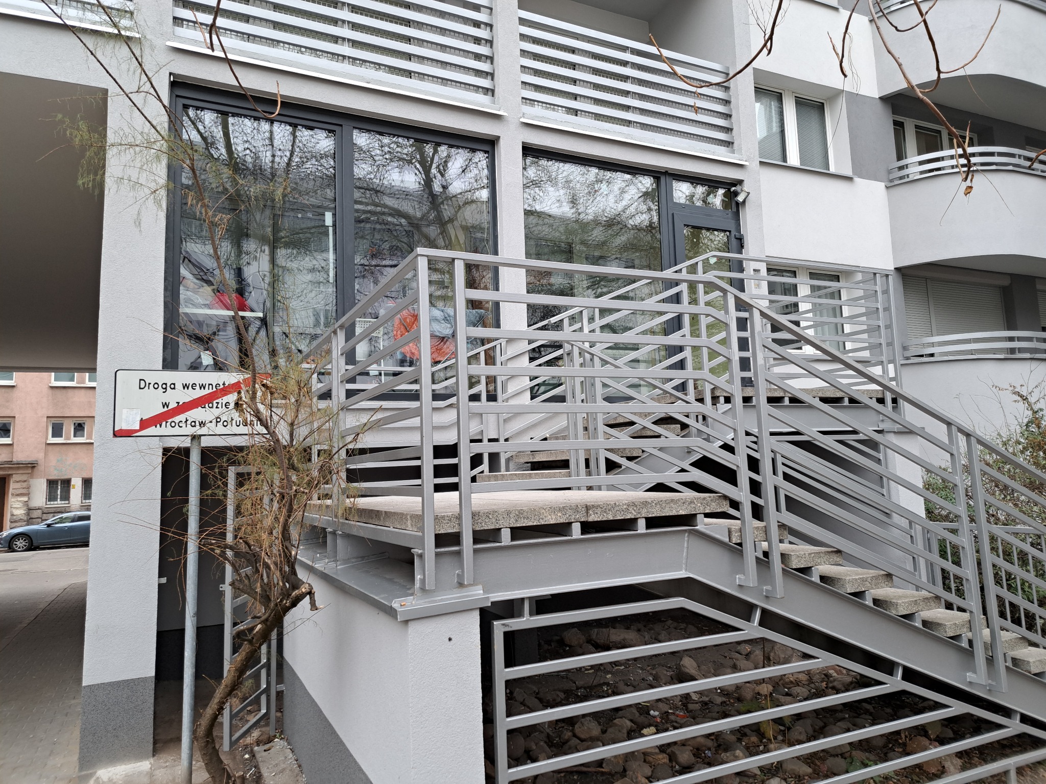 Wejście do budynku z szarymi oknami, metalową balustradą i schodami; w szybach okien odbija się otoczenie.
