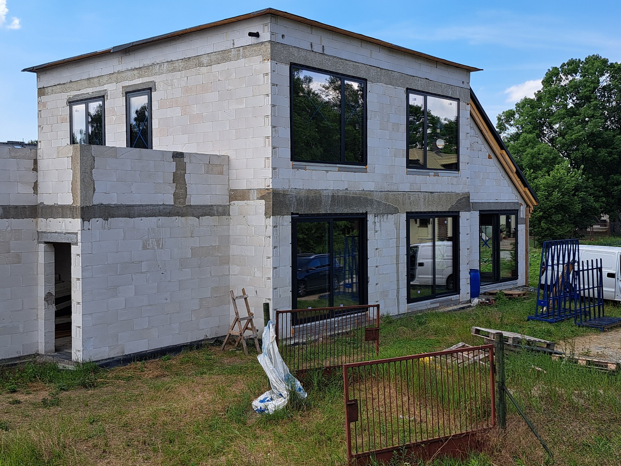 Surowy stan domu w trakcie budowy z zamontowanymi oknami z czarną ramą, widoczne rusztowania, palety i gruz na trawniku.