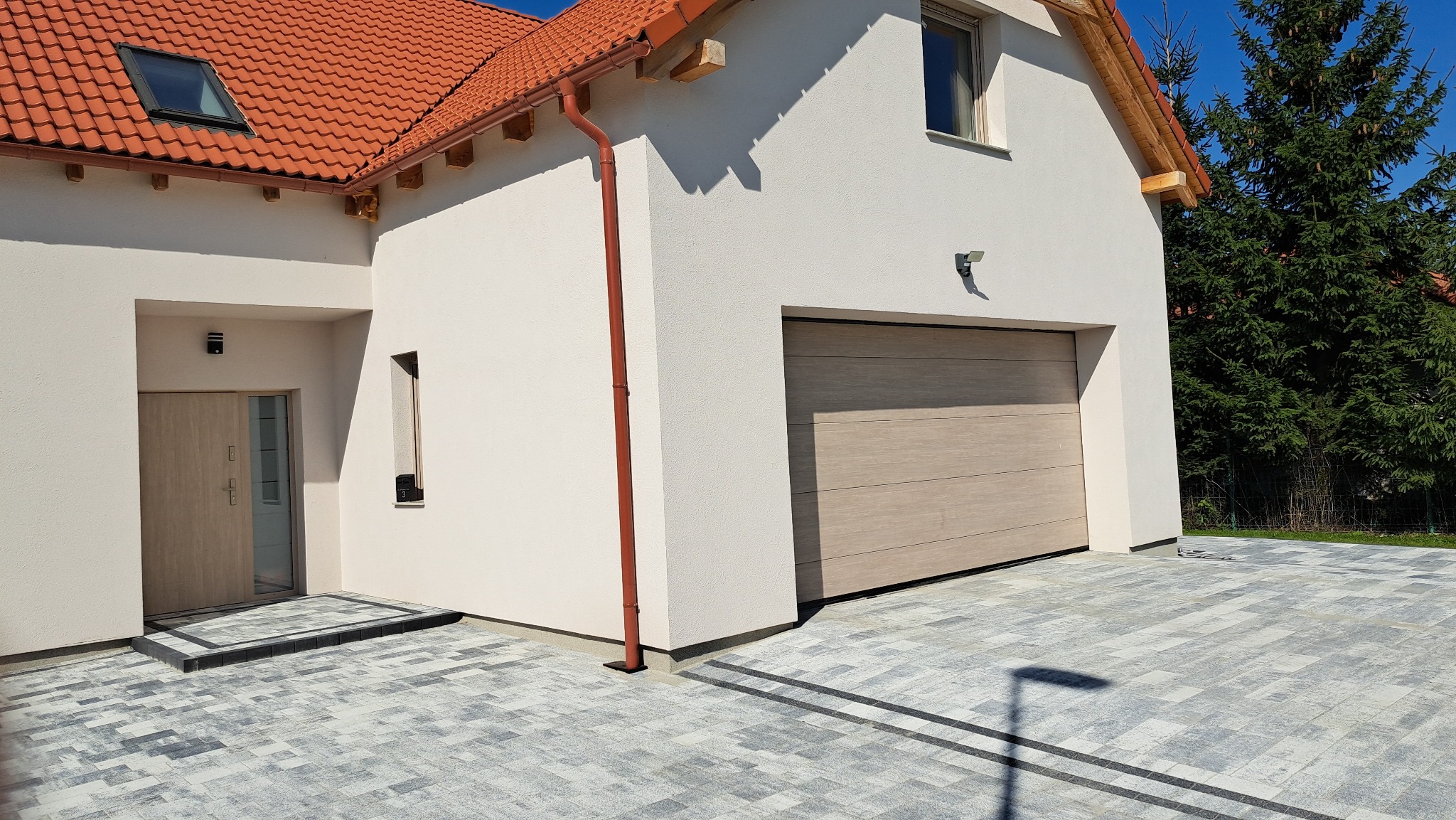 Jasna brama garażowa segmentowa z poziomymi panelami w kolorze drewna, zamontowana w białym budynku z czerwoną dachówką, widoczna z zewnątrz na tle kostki brukowej.