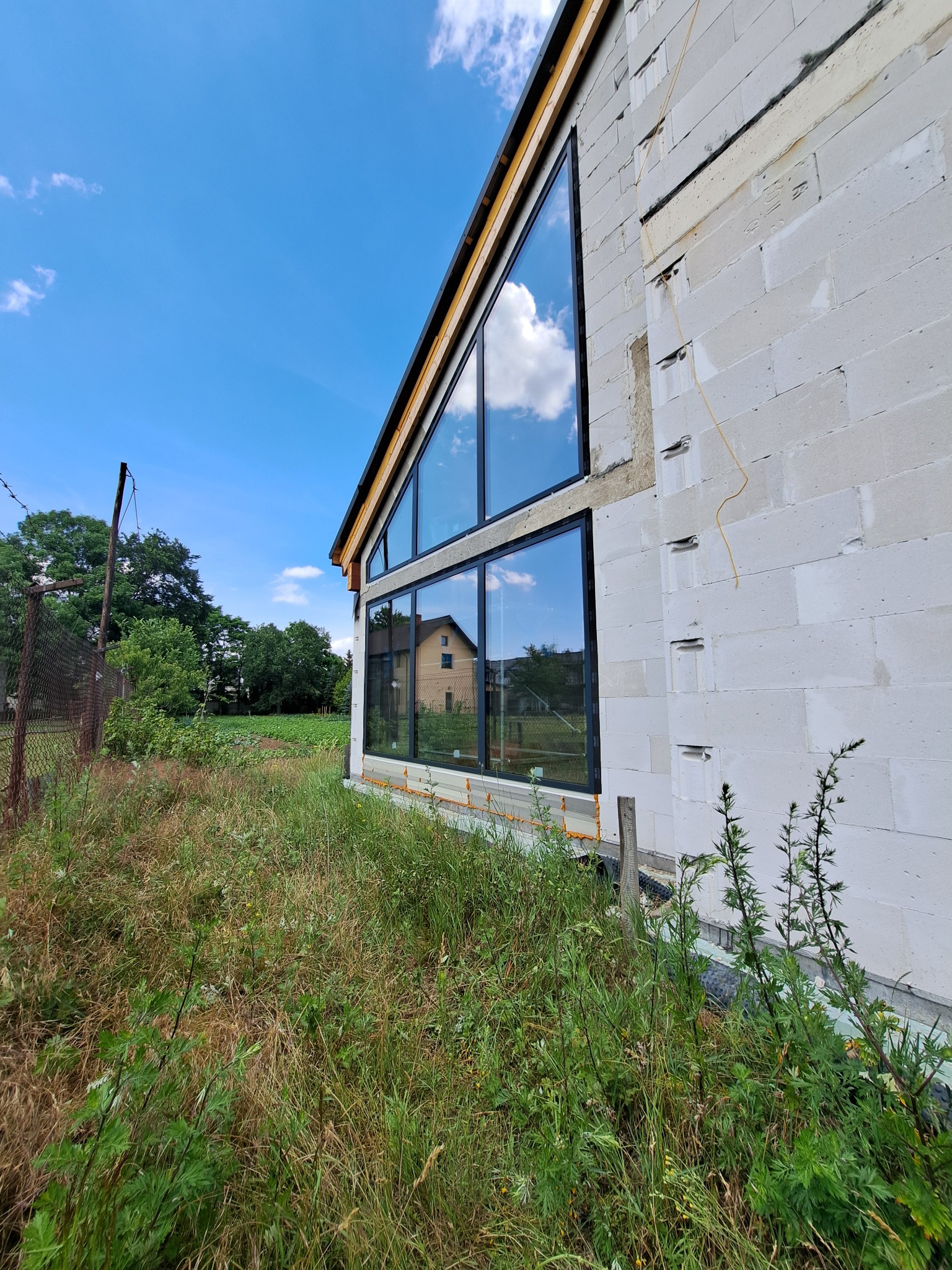 Surowy stan budynku z zamontowanymi dużymi, czarnymi oknami z widokiem na niebo i odbijającymi się chmurami, otoczony wysoką trawą i chwastami.