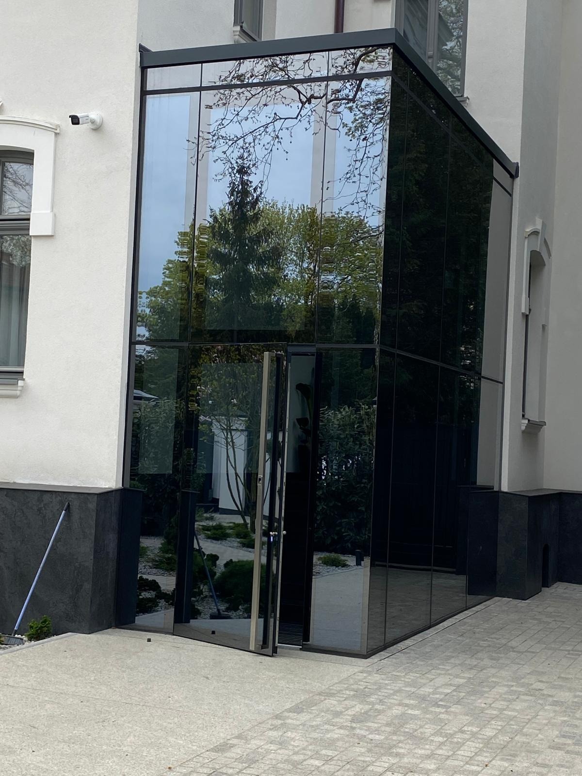 Nowoczesne przeszklone wejście do budynku z otwartymi drzwiami, odbijające zieleń ogrodu i błękit nieba.