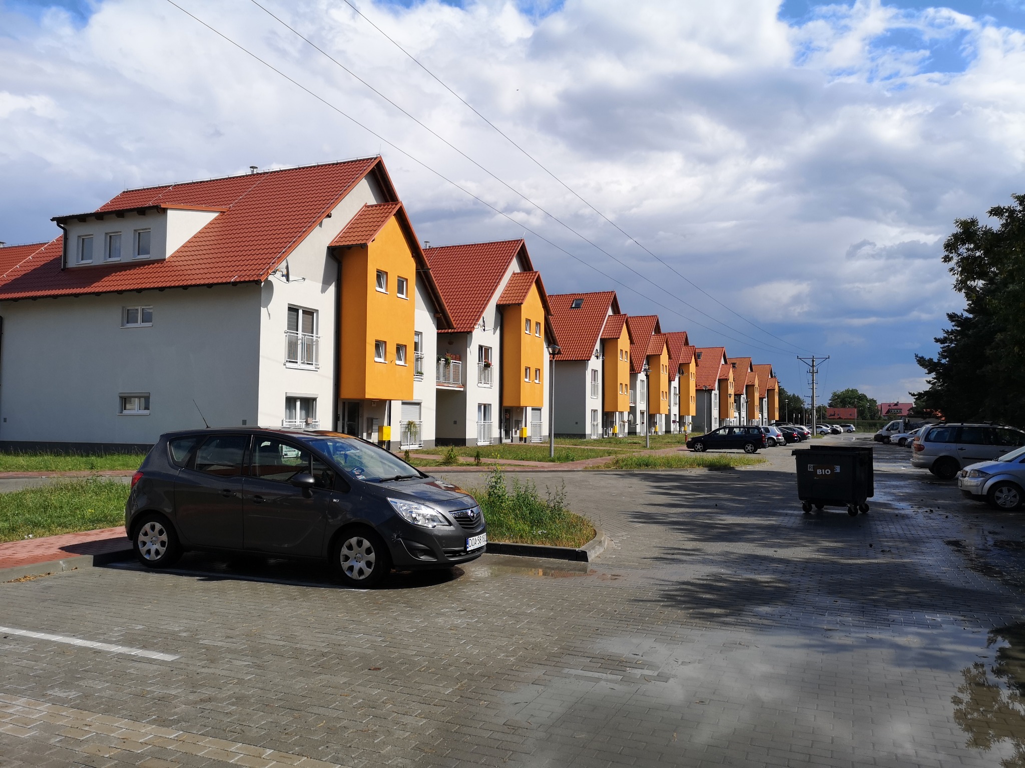 Szereg identycznych domów jednorodzinnych z pomarańczowymi elementami elewacji i czerwoną dachówką, widzianych z perspektywy ulicy z zaparkowanym samochodem i kałużami po deszczu.