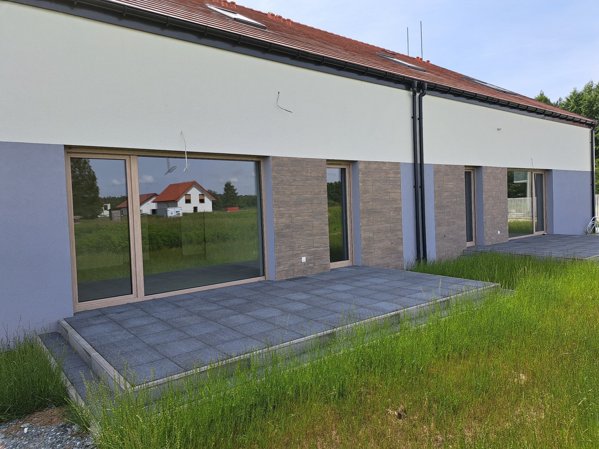 Elewacja szeregowca z dużymi oknami z drewnianą ramą i wyjściem na taras z szarej kostki, widok z poziomu trawy.