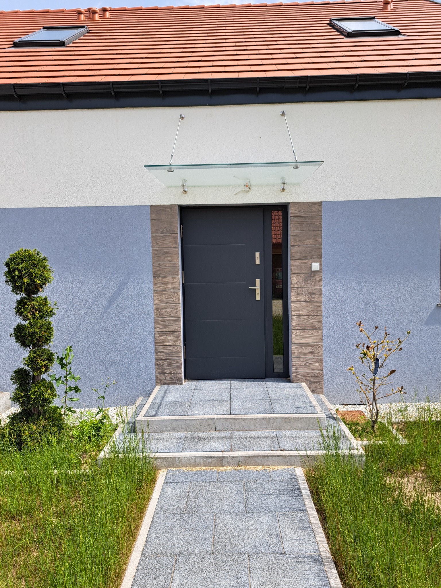 Nowoczesne, antracytowe drzwi wejściowe z przeszklonym panelem bocznym, osłonięte szklanym daszkiem, prowadzące po stopniach z szarej kostki, otoczone zielenią.