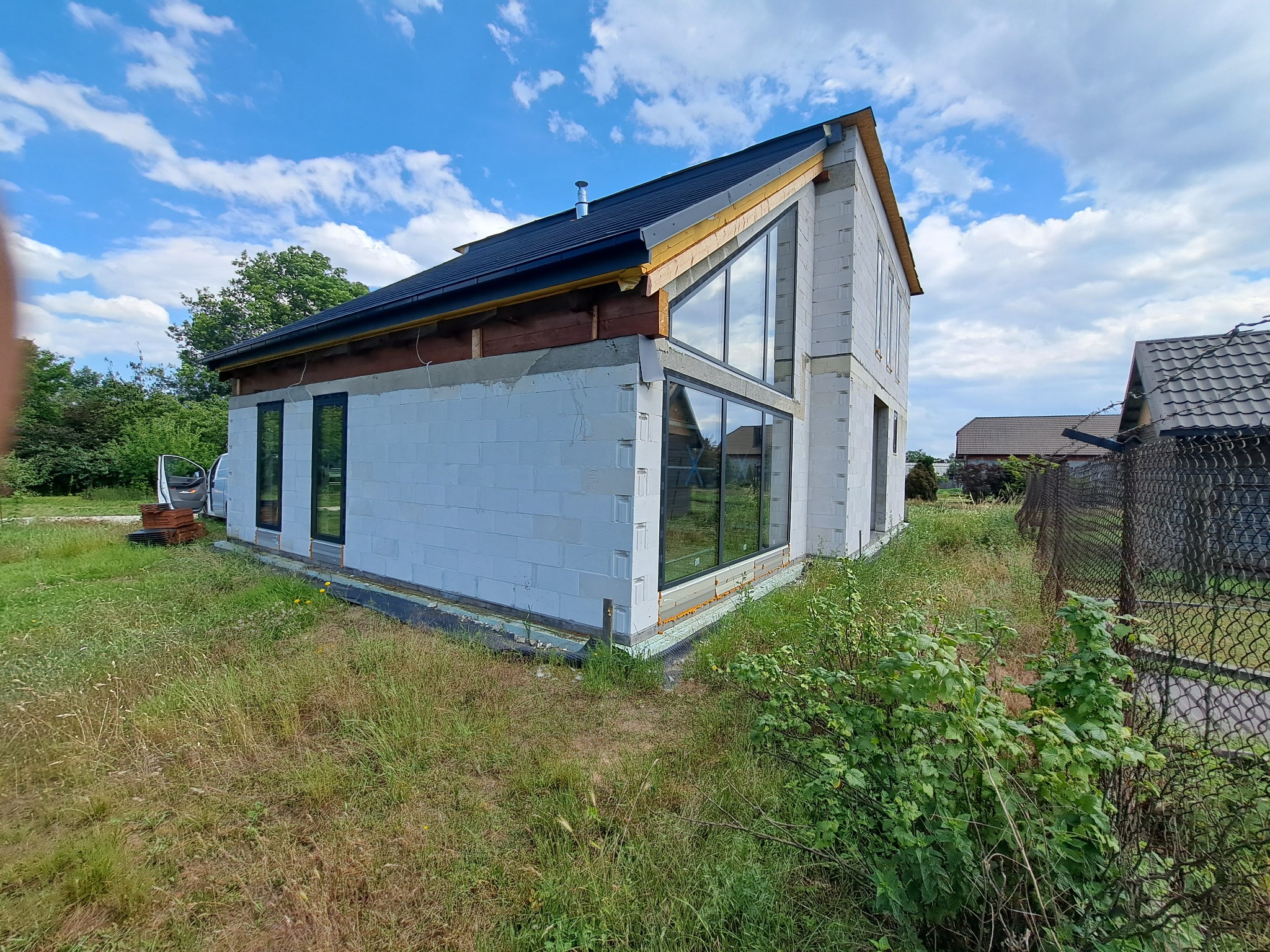 Surowy stan dwukondygnacyjnego domu z szarej cegły z zamontowanymi oknami w czarnych ramach, widoczne ocieplenie i drewniane elementy konstrukcyjne dachu, otoczenie zarośnięte trawą.