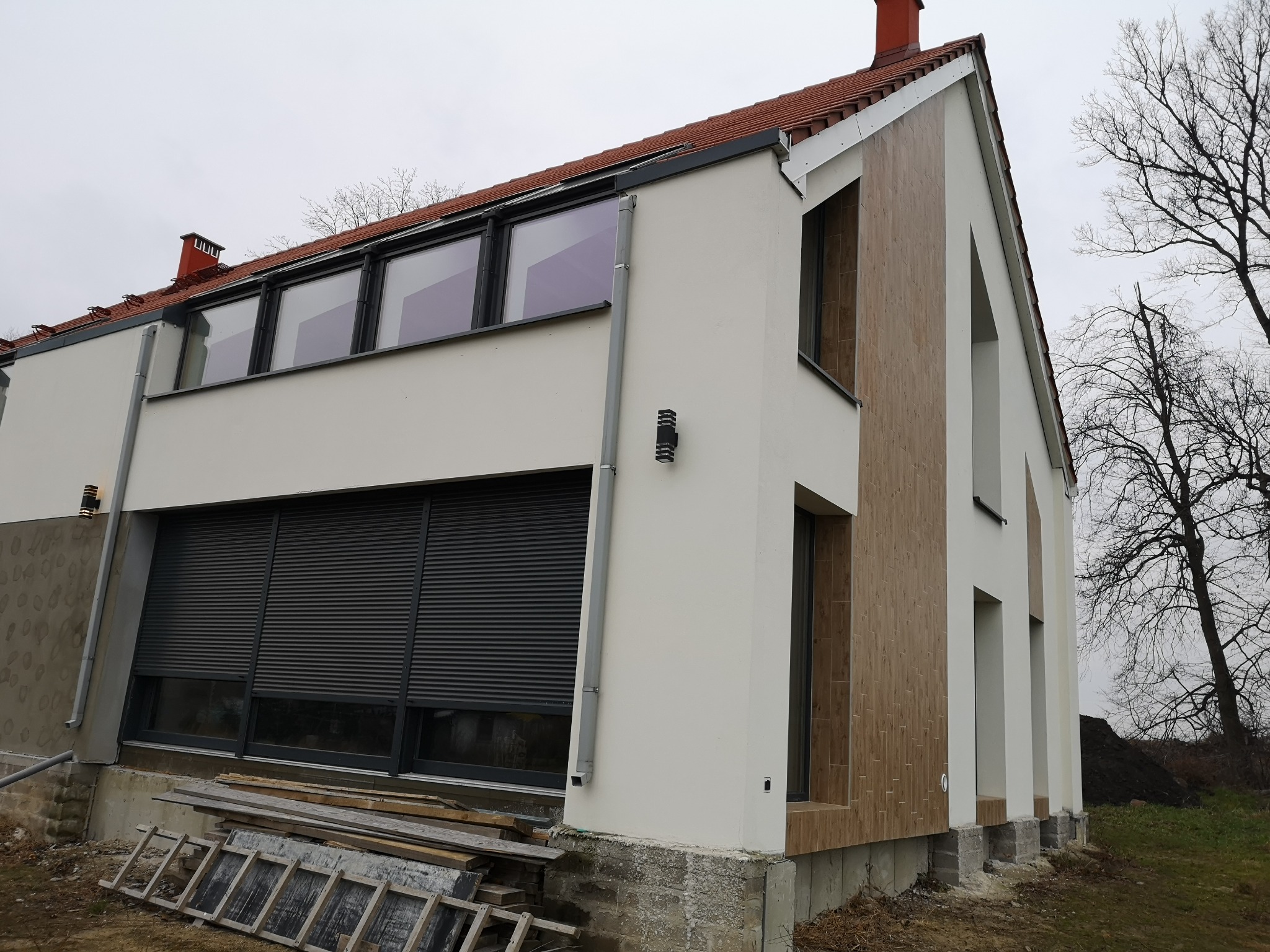 Nowoczesny dom jednorodzinny z szarymi roletami zewnętrznymi, oknami PCV i elewacją z elementami drewnopodobnymi, widoczne rusztowanie i materiały budowlane.