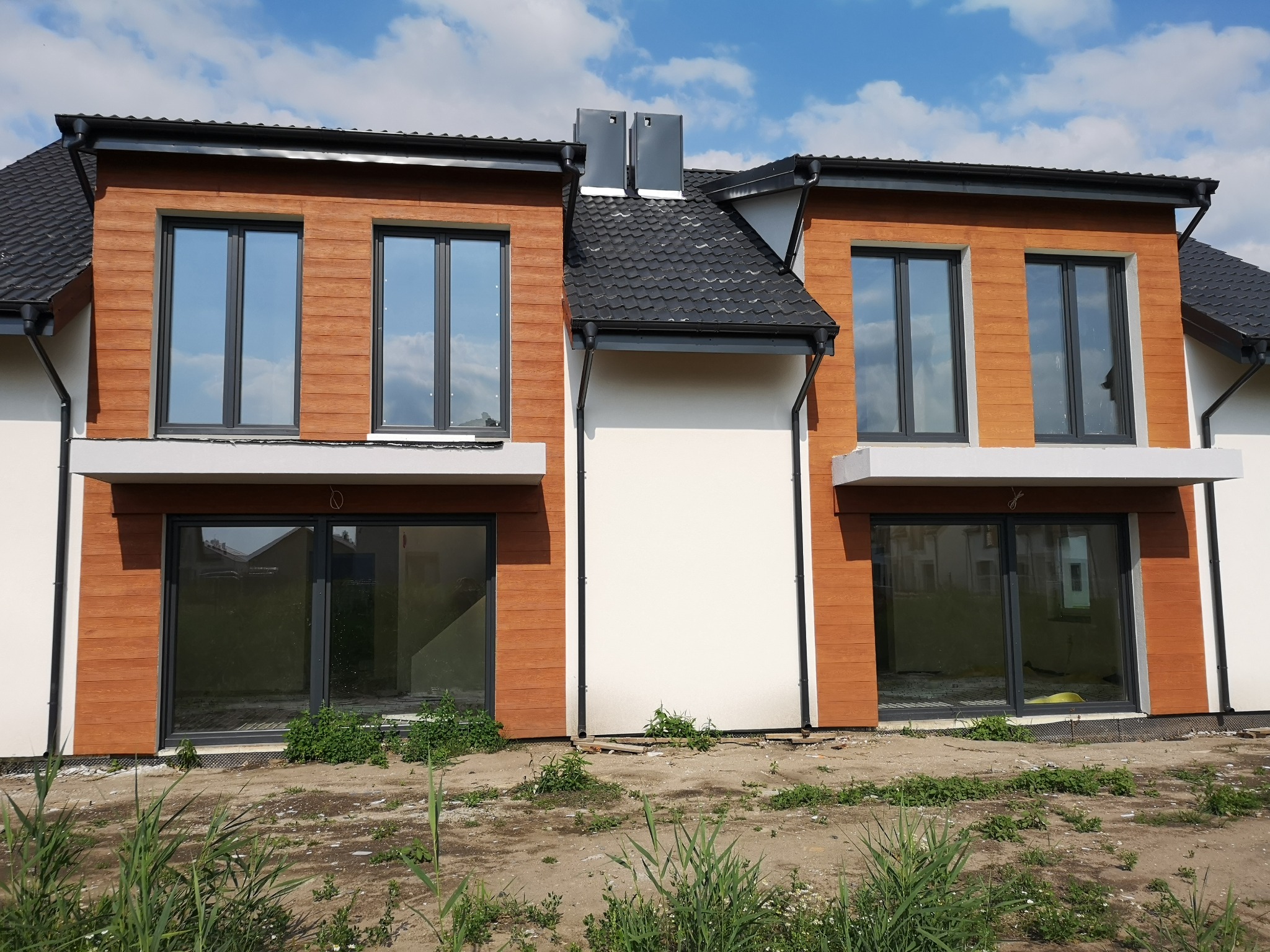 Nowoczesny dwukondygnacyjny dom w zabudowie bliźniaczej z brązową elewacją imitującą drewno, czarnymi ramami okiennymi i szarą dachówką, widziany z poziomu gruntu na etapie wykończenia terenu.