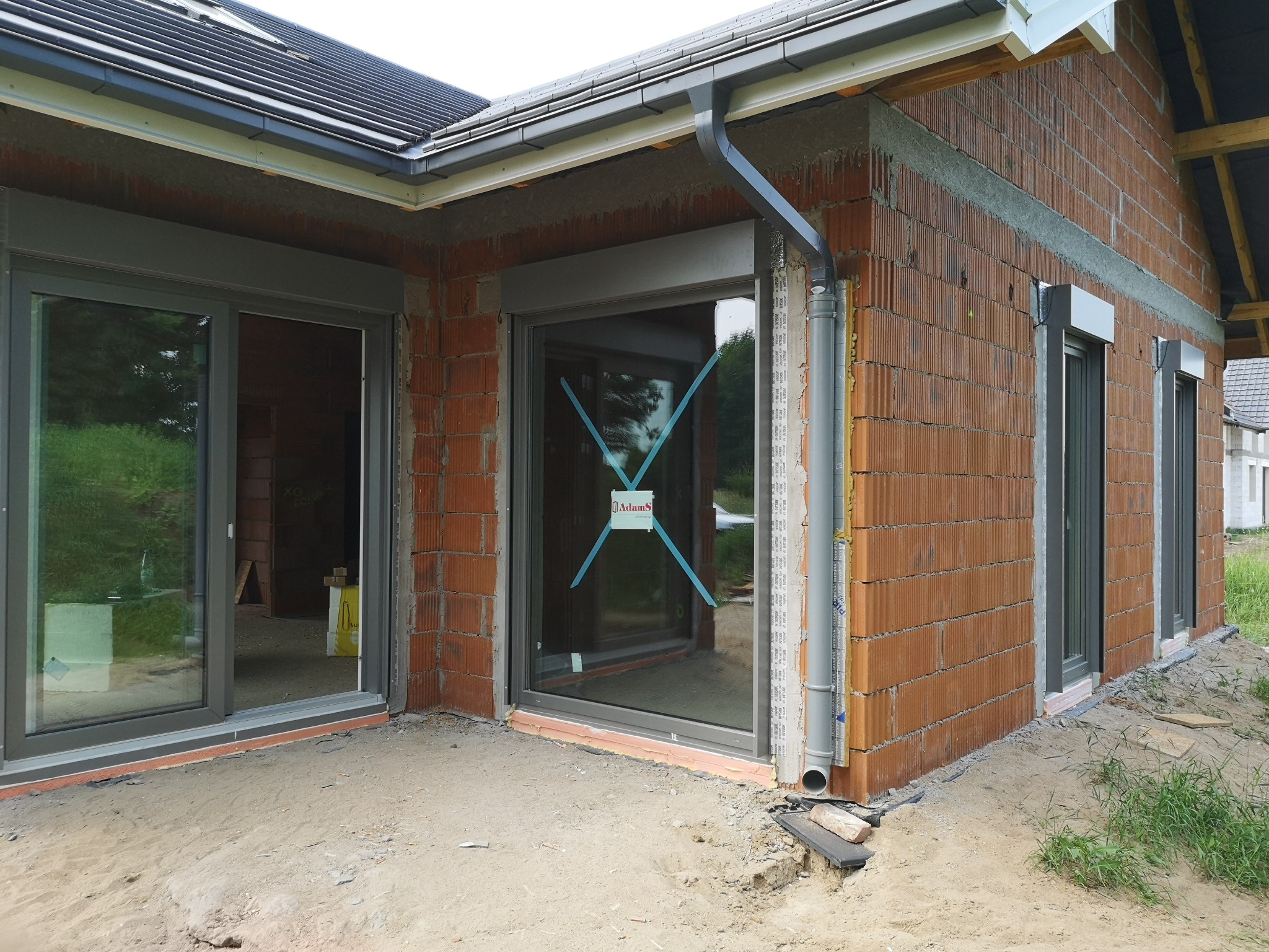 Nowo wybudowany dom w stanie surowym zamkniętym z zamontowanymi oknami w szarych ramach i częściowo opuszczonymi roletami zewnętrznymi; na szybie jednego z okien naklejka producenta.