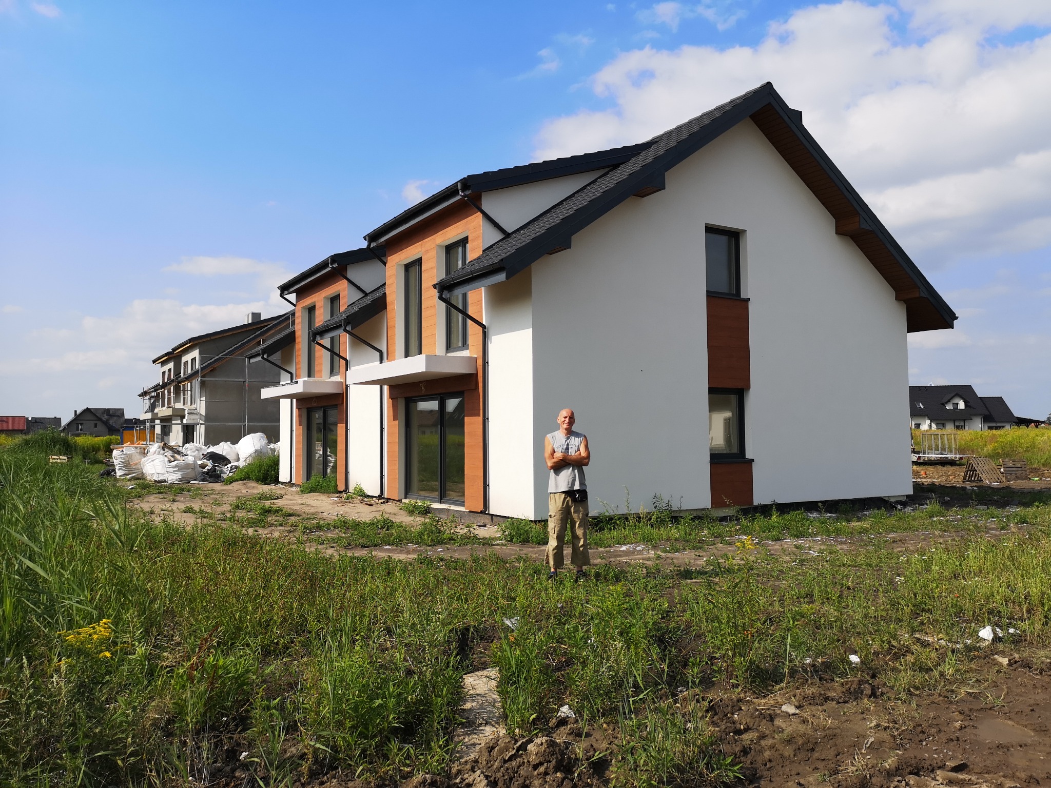 Nowoczesny dom jednorodzinny z dużymi oknami, na pierwszym planie mężczyzna z założonymi rękami, teren wokół domu jeszcze niezagospodarowany.