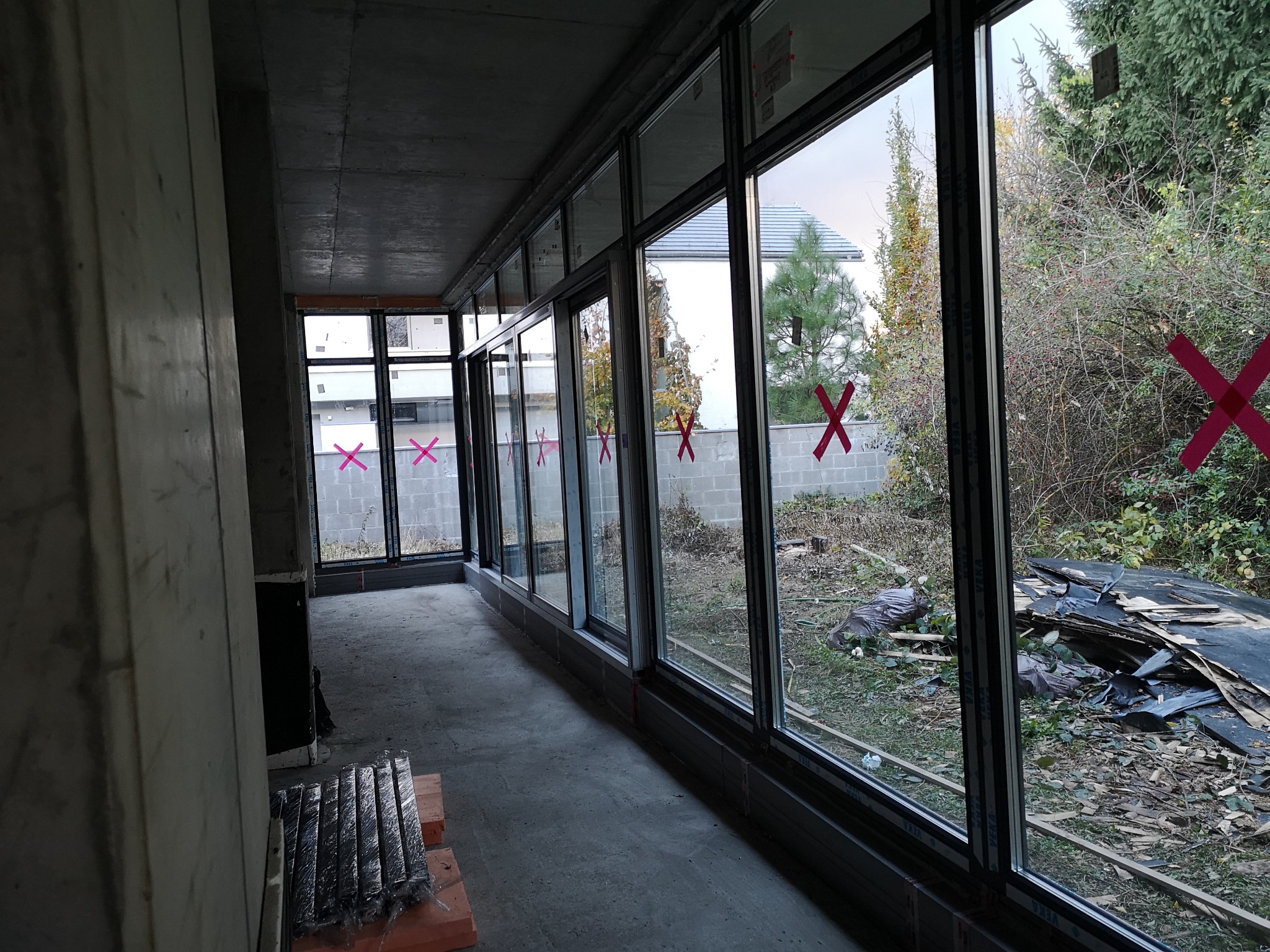 Wnętrze w trakcie montażu okien z czarnymi ramami, widoczne czerwone oznaczenia na szybach i ścianie, surowy beton oraz widok na ogród zza okien.