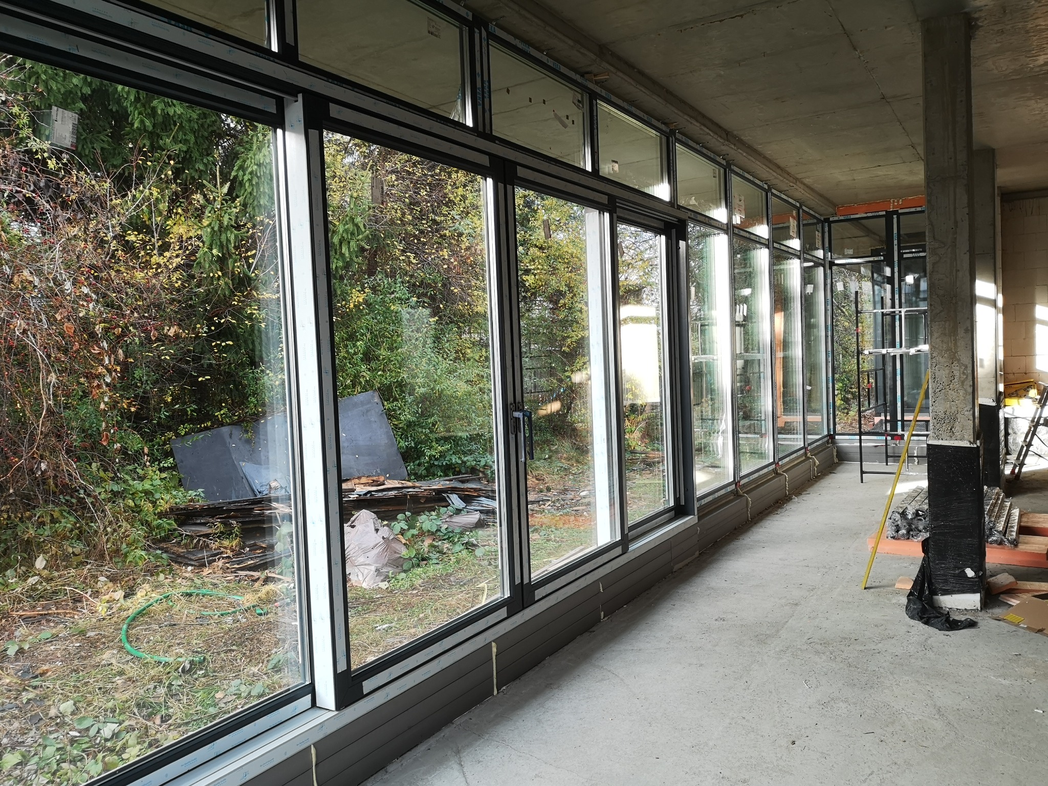 Zamontowane okna aluminiowe z czarnymi ramami w surowym stanie budynku, widok na zarośnięty ogród z elementami gruzu i węża ogrodowego.