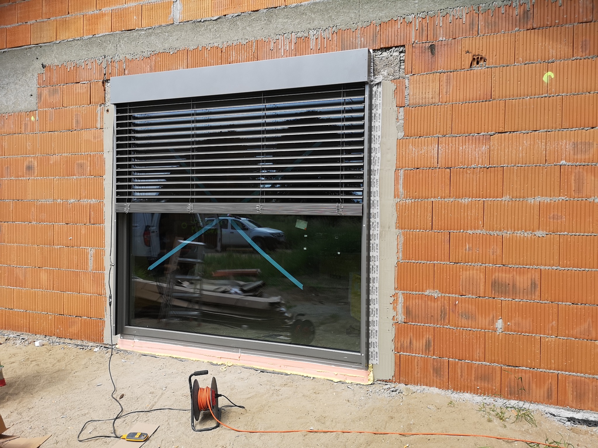 Montaż szarych żaluzji fasadowych w oknie budynku z czerwonej cegły, widoczne narzędzia i przewody elektryczne na placu budowy.