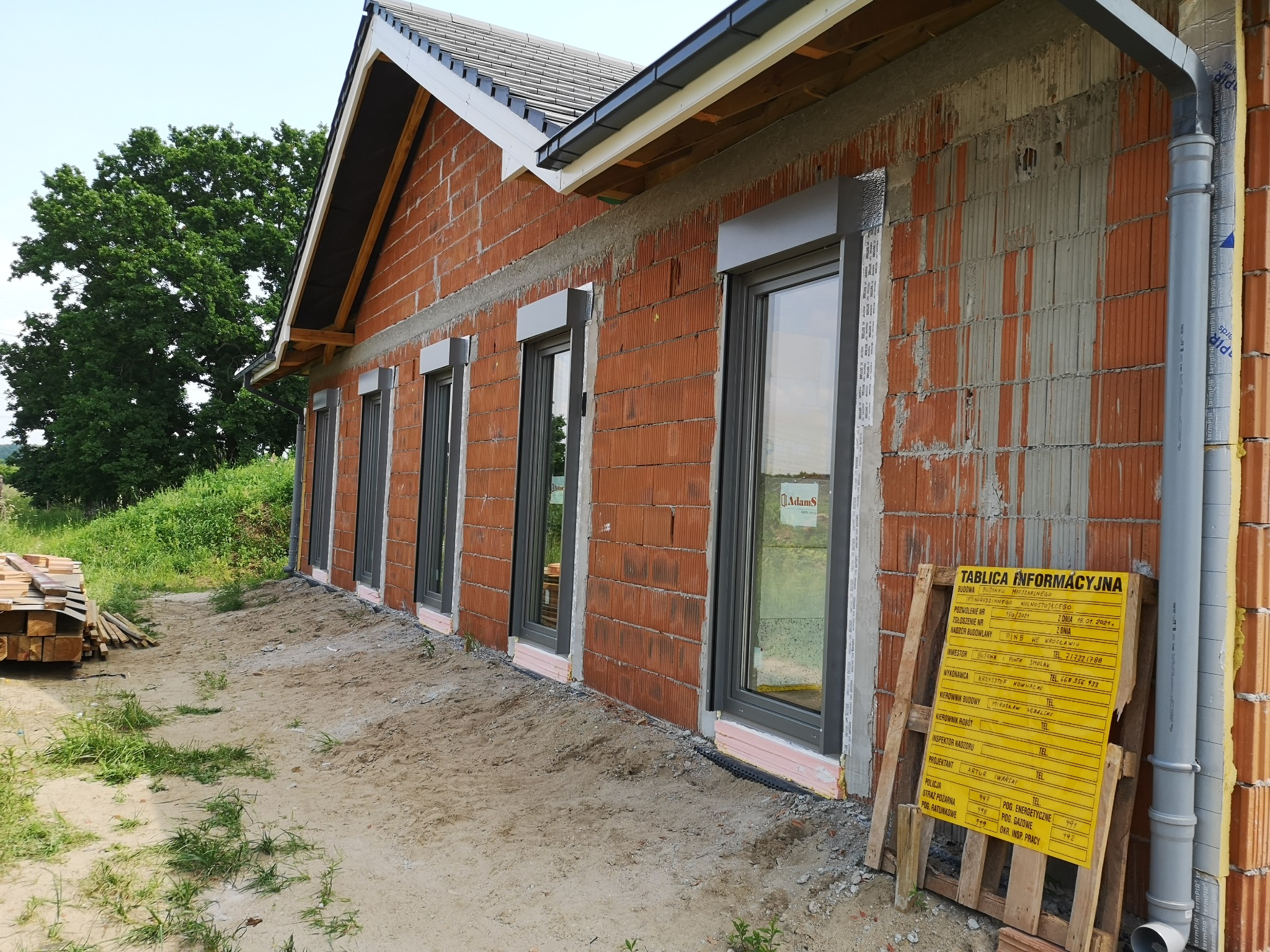 Nowo wybudowany dom z cegły z zamontowanymi oknami z szarymi ramami i zewnętrznymi roletami w kolorze szarym, widoczne stosy drewna i tablica informacyjna budowy.