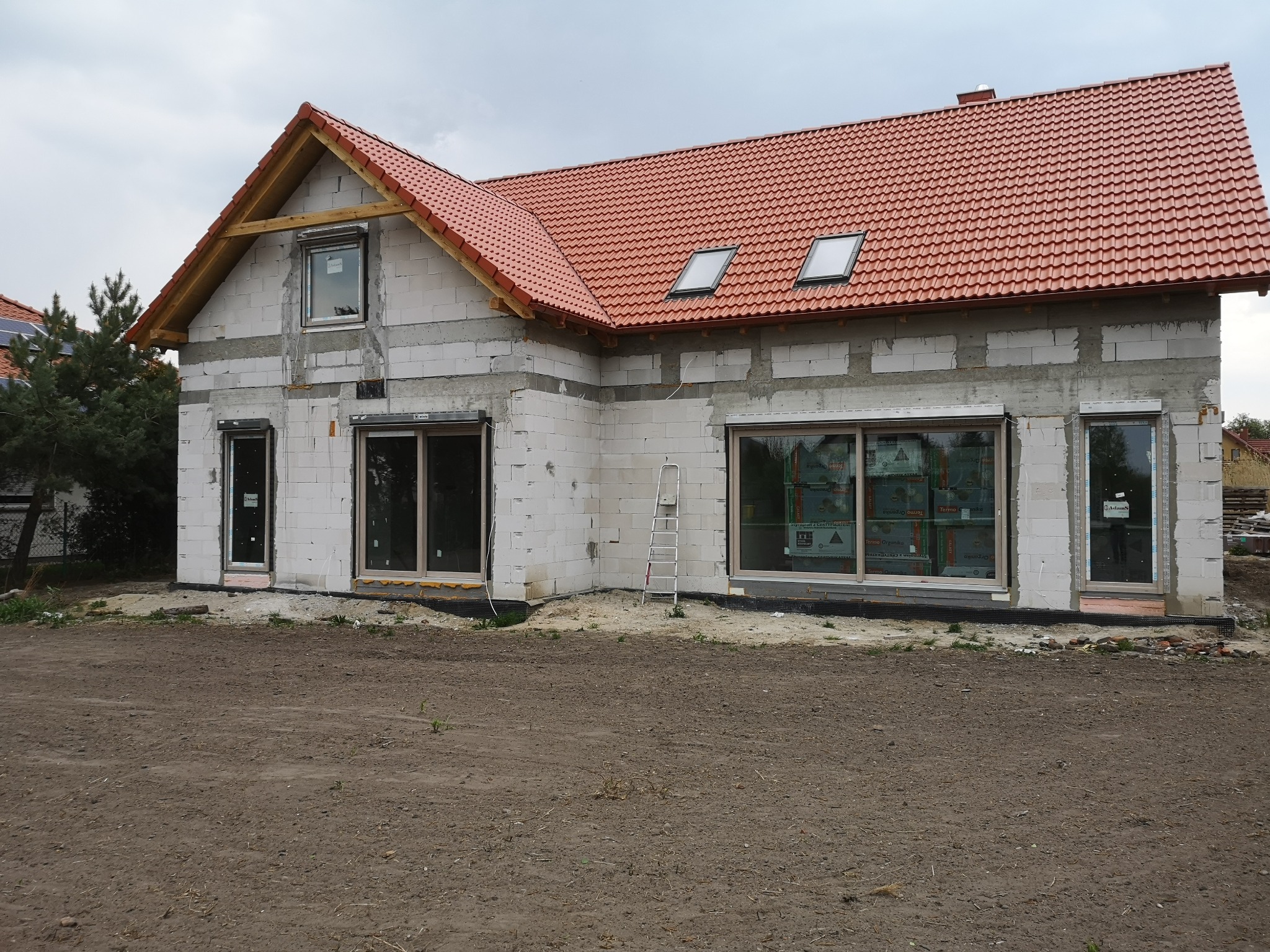 Surowy stan budynku jednorodzinnego z zamontowanymi oknami, widoczne materiały budowlane i drabina oparta o ścianę, dach pokryty czerwoną dachówką.