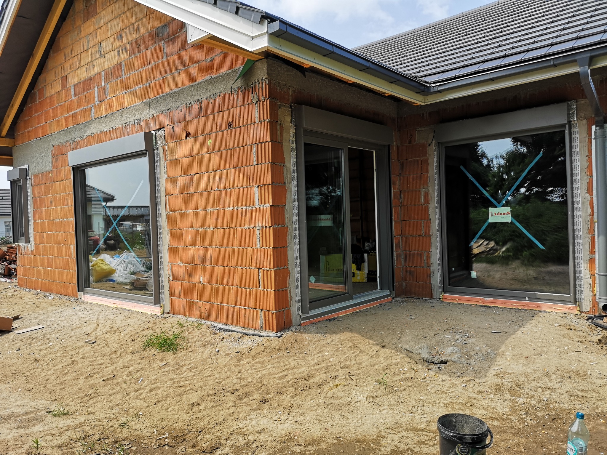 Surowy stan budynku z zamontowanymi oknami przesuwnymi z szarymi ramami i roletami zewnętrznymi; widoczne niedokończone ściany z cegły, piasek na zewnątrz i niebieska taśma zabezpieczająca na szybach.