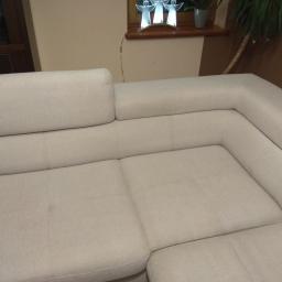 Kamil Dyczka - Jasna, narożna kanapa z tapicerką z widocznymi przeszyciami i zagłówkiem, ustawiona w salonie z dekoracyjną lampą aniołów i rośliną doniczkową w tle.