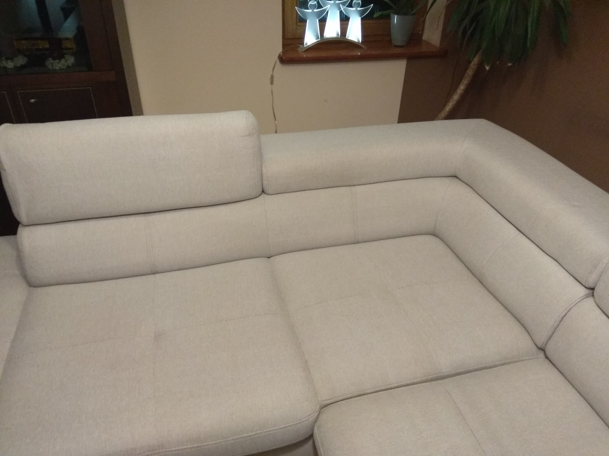 Jasna, narożna kanapa z tapicerką z widocznymi przeszyciami i zagłówkiem, ustawiona w salonie z dekoracyjną lampą aniołów i rośliną doniczkową w tle.