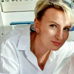 Portret kobiety w białej koszuli z kr&oacute;tkimi blond włosami, noszącej delikatną biżuterię i kolczyki w kształcie łezki, z niebieskim elementem w tle.